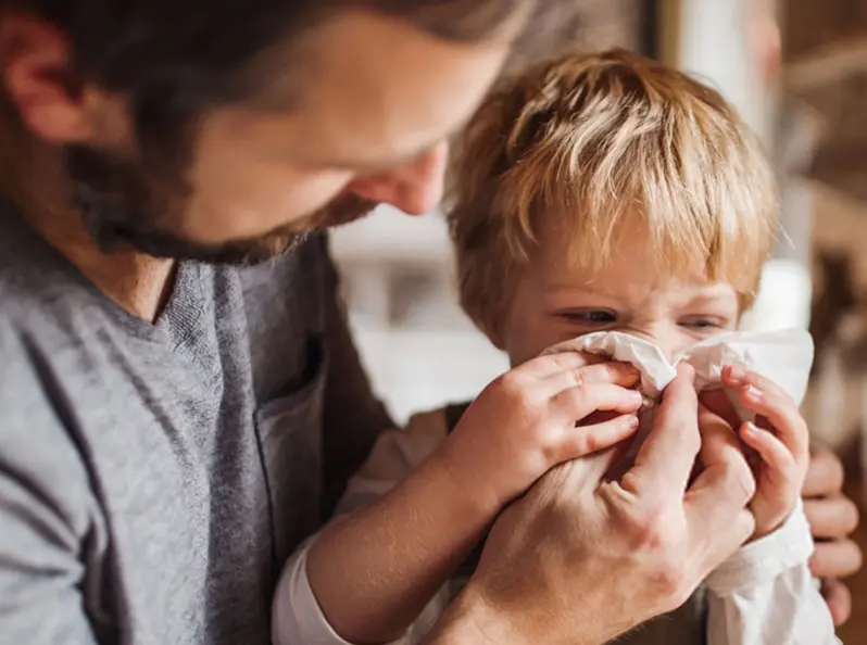 A parent helping a young child blow their nose into a tissue.