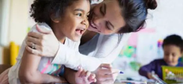 Parent leaning round to hug their smiling child. Another child is drawing in the background.