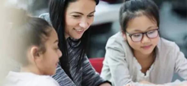 Teacher sat at a table between two students smiling and pointing at a computer screen.