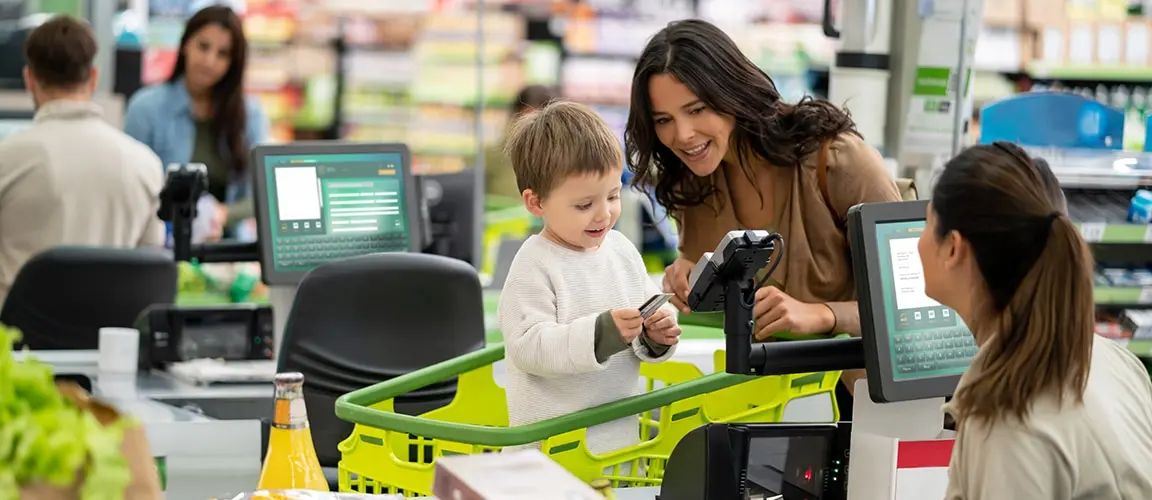 Parent showing child how to insert credit card into card reader at grocery store checkout. The Cashier is smiling at them